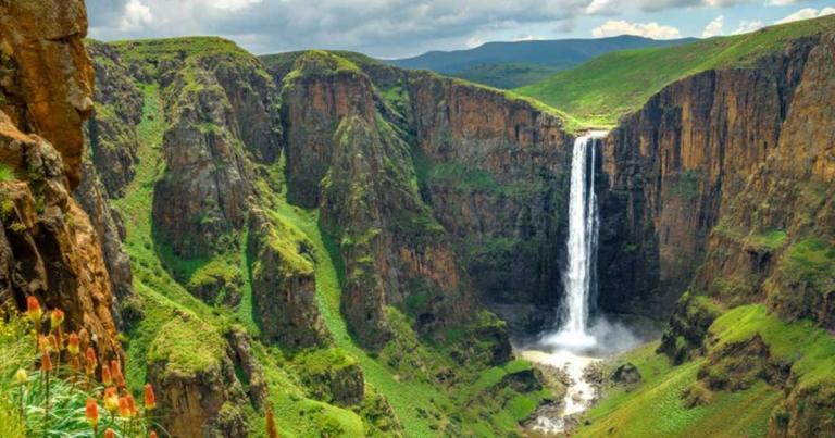 A tall waterfall cascades down a steep cliff surrounded by lush green hills under a cloudy sky.