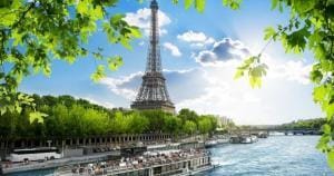 The Eiffel Tower by the Seine River on a sunny day, framed by green leaves and a tour boat passing below.