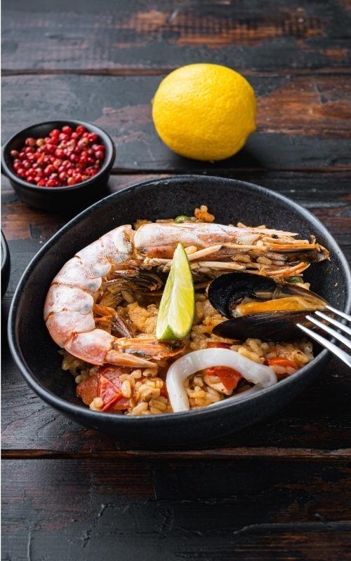 A plate of seafood paella with shrimp, mussels, squid, and a lime wedge. A lemon and a bowl of red peppercorns are in the background on a wooden table.