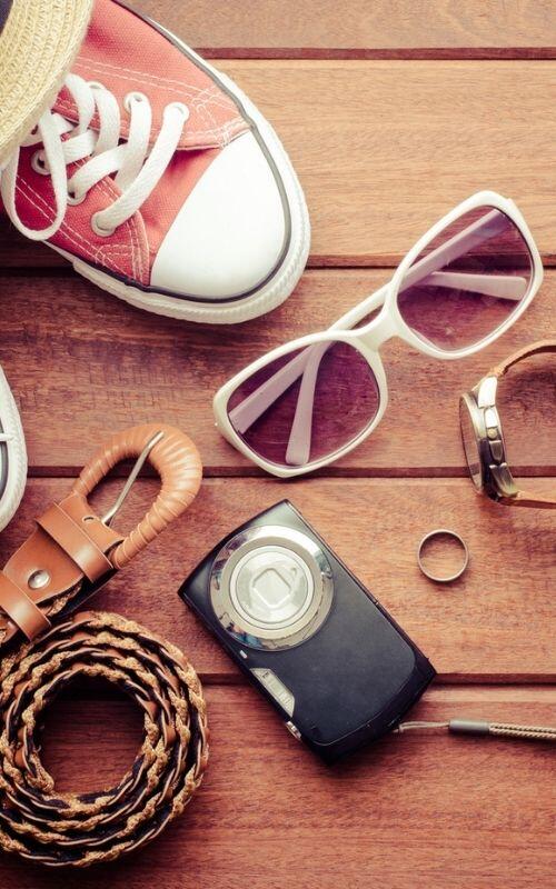 A pair of red sneakers, white sunglasses, a hat, a leather belt, watch, ring, and a vintage camera are laid out on a wooden surface.