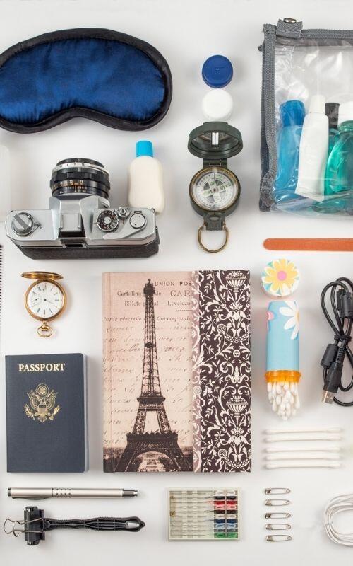 Assorted travel essentials are arranged neatly, featuring a camera, passport, compass, notebooks with Eiffel Tower designs, toiletries, watch, sewing kit, and cables on a pristine white background.