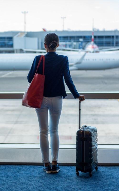 Person standing at airport window, gazing at the plane with a suitcase and tote bag.