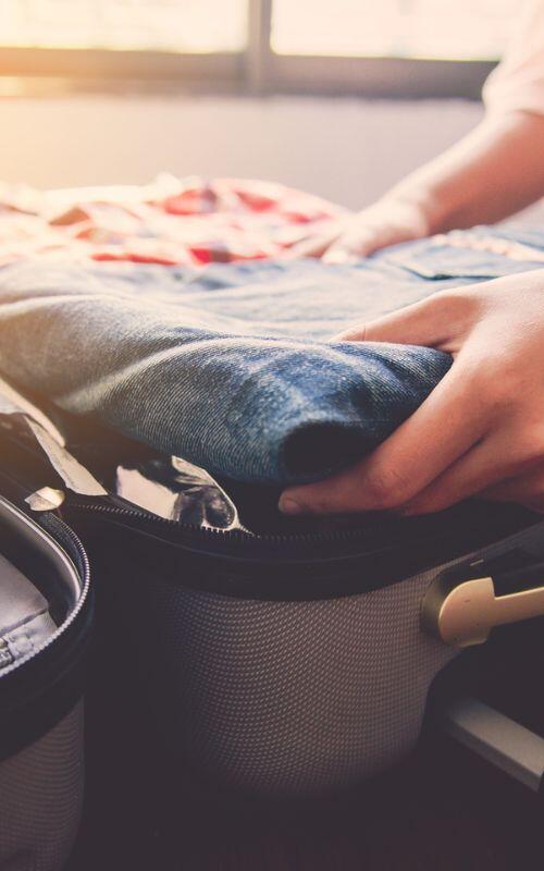 Sunlight streams through the window as someone thoughtfully packs travel essentials, including jeans, into their suitcase.