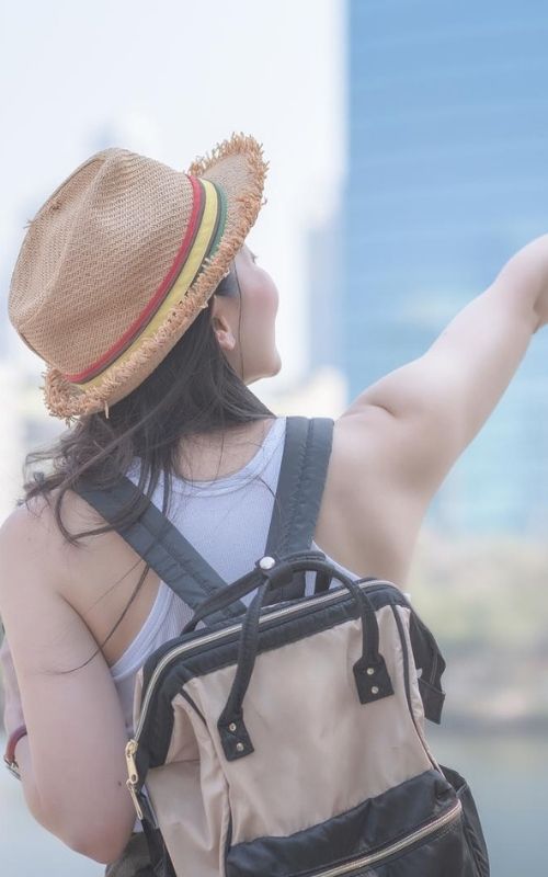 A person wearing a hat and backpack, showcasing their travel essentials, points towards a blurred cityscape background.