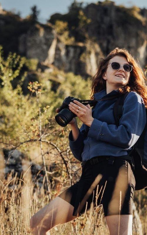 A woman with sunglasses, carrying travel essentials like a camera, stands in a grassy field with rocky cliffs in the background.