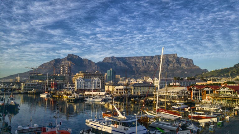 Scenic view of a coastal city with a bustling harbor, boats docked, and a mountainous backdrop under a partly cloudy sky.