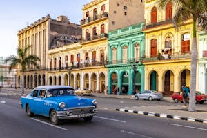 A vintage blue car drives past colorful colonial buildings on a city street lined with palm trees.