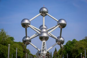 The image shows the Atomium in Brussels, a large metallic structure resembling an iron crystal, composed of connected spheres. It stands against a clear blue sky with trees surrounding the base.