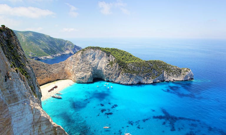 Aerial view of a bright turquoise cove with a sandy beach, surrounded by steep cliffs and lush greenery. Several boats are anchored near the shore.