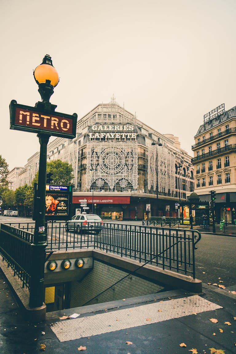 Entrance to a Paris metro station near the Galeries Lafayette, with decorative building lights and a street scene including traffic and pedestrians on a cloudy day.