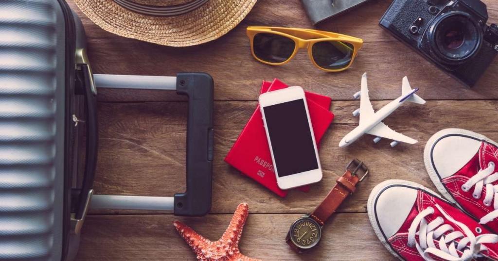 Travel essentials laid out: suitcase, hat, sunglasses, camera, phone on passports, toy airplane, red sneakers, wristwatch, and a starfish on a wooden surface.