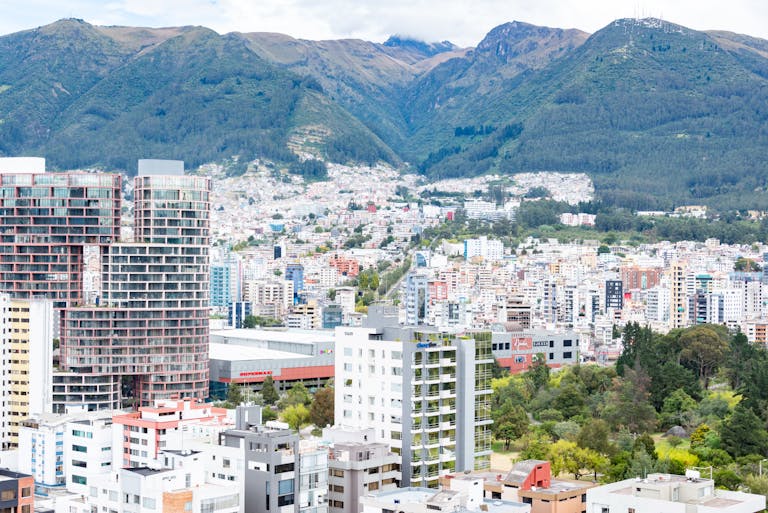 Cityscape with modern high-rise buildings in the foreground, surrounded by lush greenery and mountains in the background under a cloudy sky.