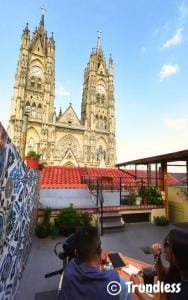Two people sit on a rooftop patio with decorative tiles, overlooking a large, ornate, twin-spired cathedral under a blue sky. Photo by trundless.com