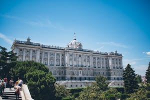 The image shows the Royal Palace of Madrid surrounded by trees, with people walking on a pathway in the foreground under a clear blue sky.