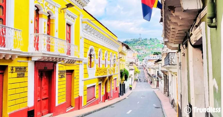 A vibrant street in the historic town of Quito features colorful yellow and red buildings with ornate balconies. A flag is visible hanging from a building on the right, and rolling hills with lush greenery can be seen in the distance under a partly cloudy sky.