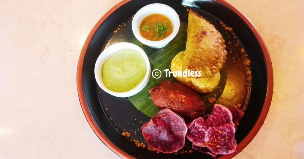 A plate with a stuffed pastry, purple sweet potato chips, chutneys, and a brown fried item on a banana leaf.