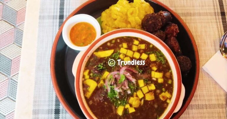 A plate with soup, crispy elements, fried plantain, sausage, and a side of sauce on a textured tablecloth.