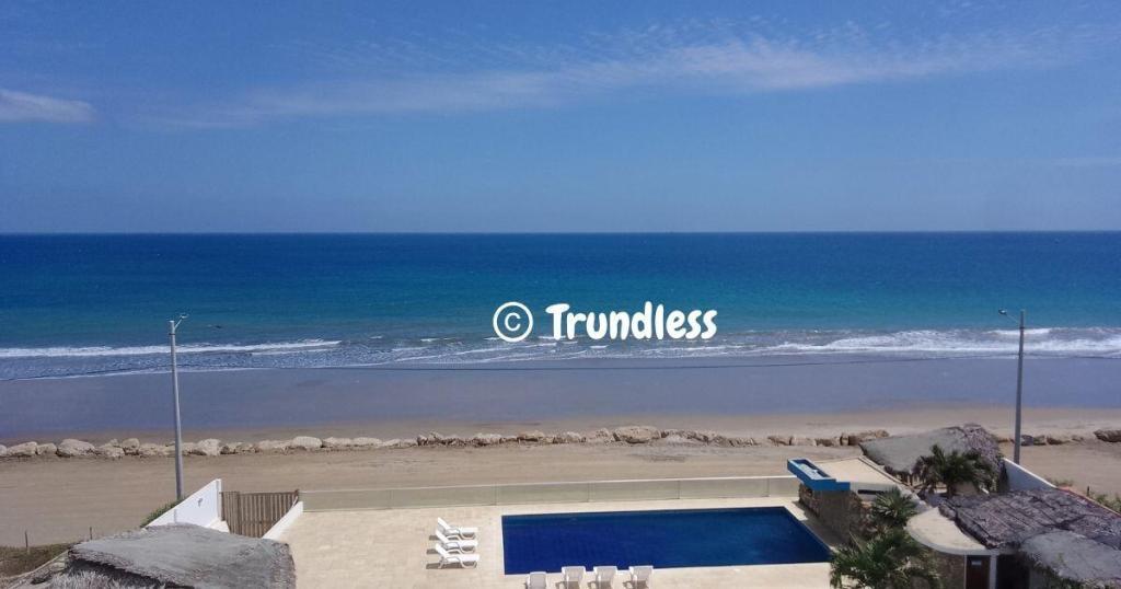 A beachfront view with a clear blue ocean, sandy shore, and a swimming pool in the foreground offers an idyllic setting for those considering permanent residence in Ecuador. A few white lounge chairs are perfectly arranged by the pool to enjoy this tropical paradise every day.