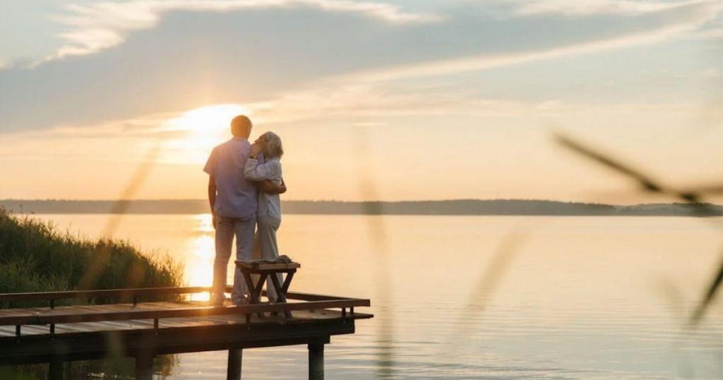 A couple stands on a wooden dock embracing, facing a serene sunset over a calm lake with soft clouds in the sky.