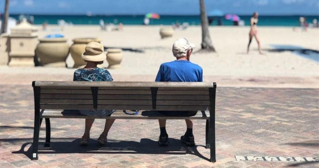 Two people wearing hats sit on a bench facing a sandy beach with a few people and umbrellas in the background.