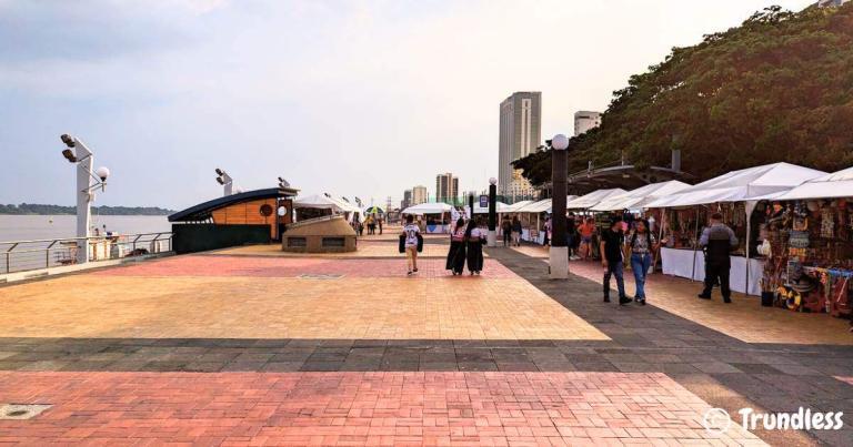 People walking along a riverside promenade lined with vendor stalls and trees. Urban buildings are visible in the background.