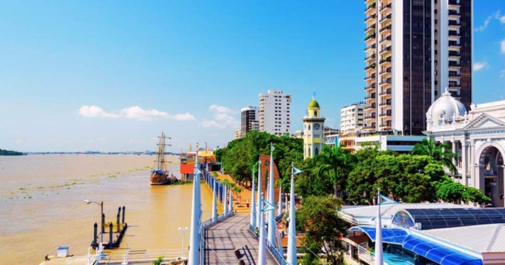 Riverfront view with a promenade, tall buildings, a yellow clock tower, and a ship on the river under a clear blue sky.