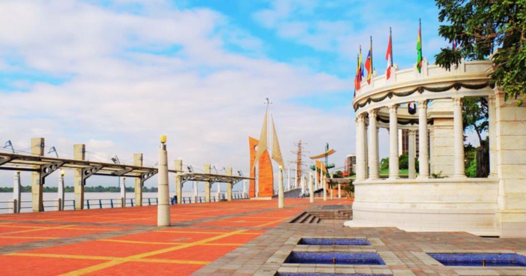 A riverside promenade with flag poles, a circular pavilion, and modern sculptures along a red and gray paved path under a partly cloudy sky.