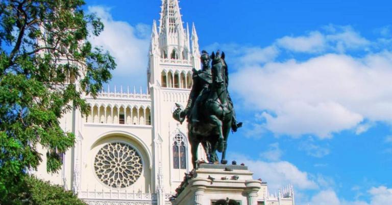 Equestrian statue in front of a Gothic-style cathedral with a large rose window, framed by trees, under a clear blue sky.