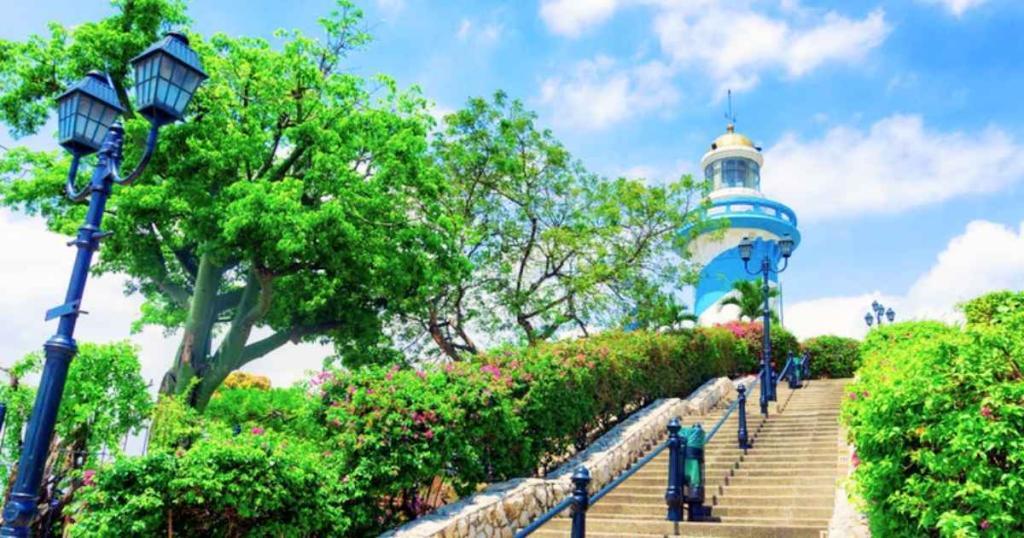 A stone staircase leads up to a blue lighthouse, surrounded by lush green trees and colorful flowers under a bright blue sky.