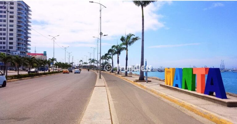 A coastal road lined with palm trees near large colorful letters spelling "MANTA," with buildings on the left and the sea on the right under a partly cloudy sky.