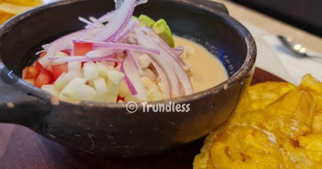 A bowl of soup with sliced red onions, diced tomatoes, and avocado, accompanied by fried plantains on the side.