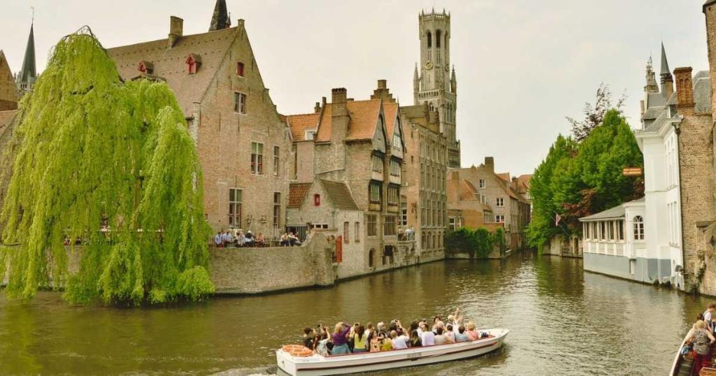 A tour boat travels along a canal in Bruges, Belgium, bordered by historic brick buildings and lush greenery, with a tall belfry visible in the background.