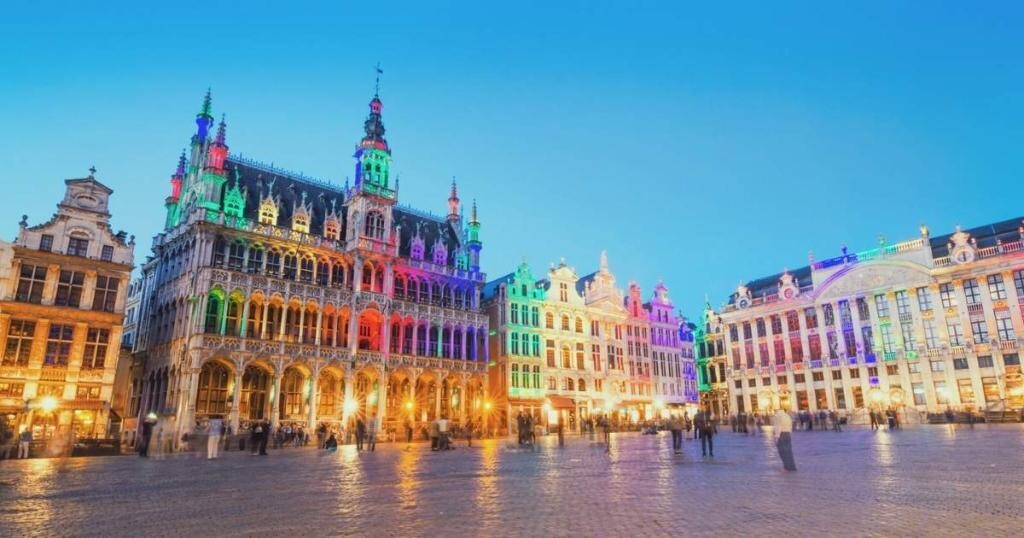 The Grand Place in Brussels illuminated with colorful lights at twilight, showcasing historic buildings and a clear sky.