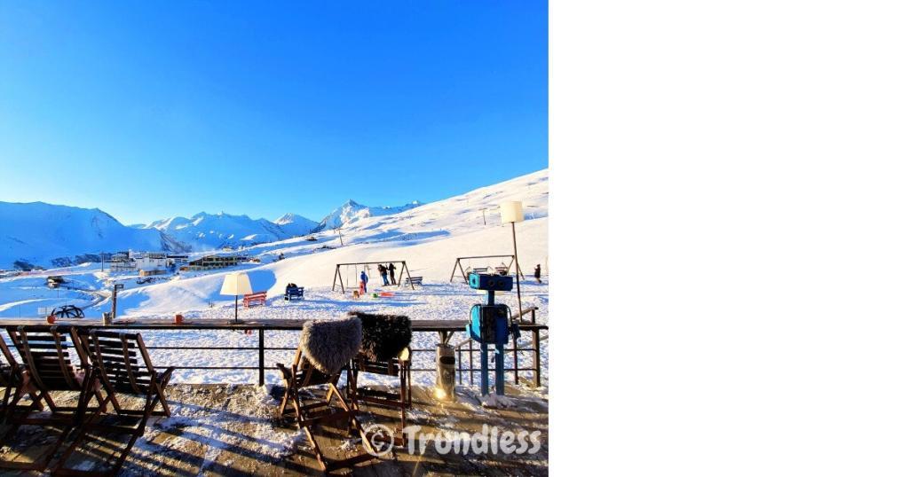 Ski resort with snowy slopes, people skiing, and chairs on a wooden deck under a clear blue sky.