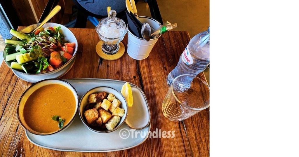 A table with a salad, soup with croutons, dessert jar, utensils in a container, a water bottle, and a glass.