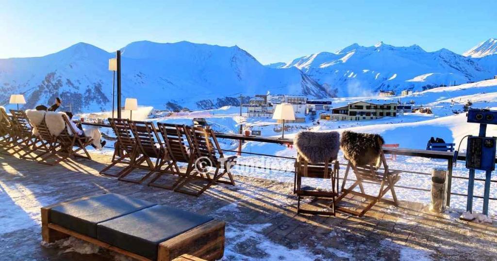 Scenic snowy mountain view from a terrace with wooden chairs and fur throws, overlooking a distant resort.