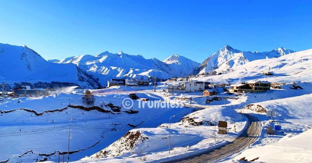 Snow-covered mountainous landscape with scattered buildings and a curving road under a clear blue sky.