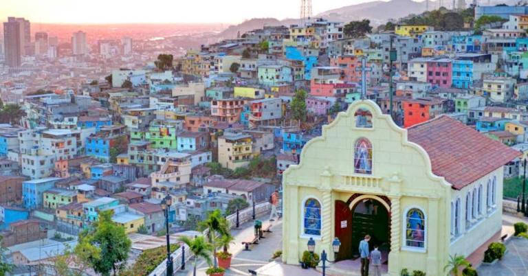 A yellow church with a red roof overlooks a colorful hillside neighborhood with numerous multicolored houses, under a setting sun.