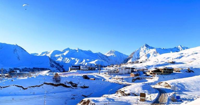 Snow-covered mountain landscape with scattered buildings and a paraglider in the sky. Bright blue sky in the background.