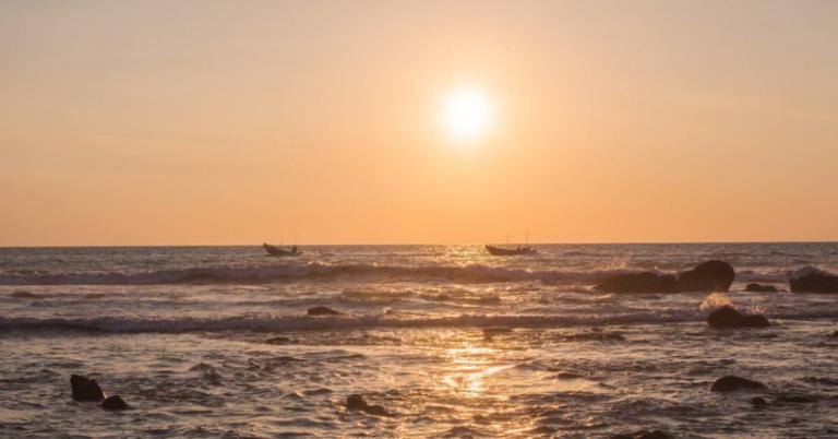 Sunset over the ocean with two boats on the horizon and waves gently crashing on rocks in the foreground.