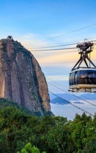 A cable car travels towards the top of a cliff with a station on it, surrounded by lush greenery and overlooking the ocean.