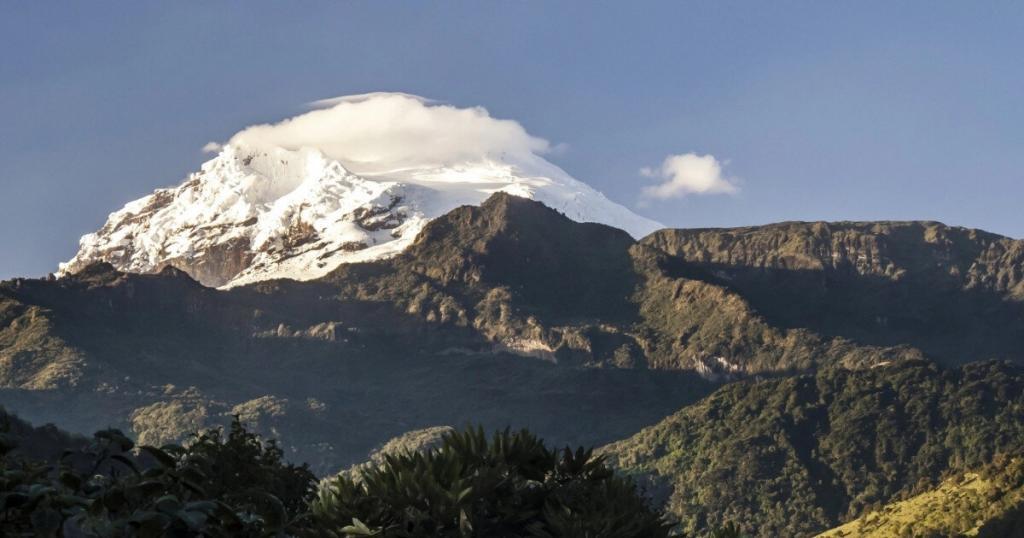 A snow-capped mountain under a clear blue sky, with a layer of clouds near the peak and lush green hills in the foreground.