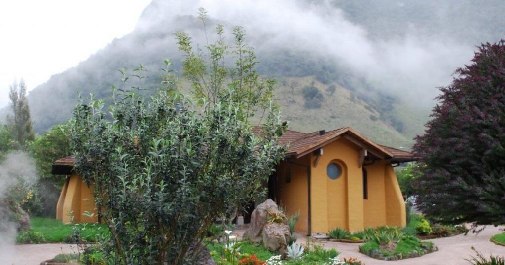 A round, earth-toned house with circular windows is set in a lush garden. Misty hills rise in the background, partially obscured by clouds.