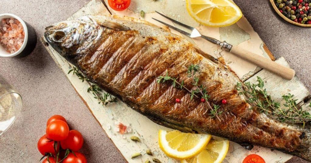 Grilled fish garnished with herbs and pepper on a cutting board, surrounded by lemon slices, tomatoes, and spices.