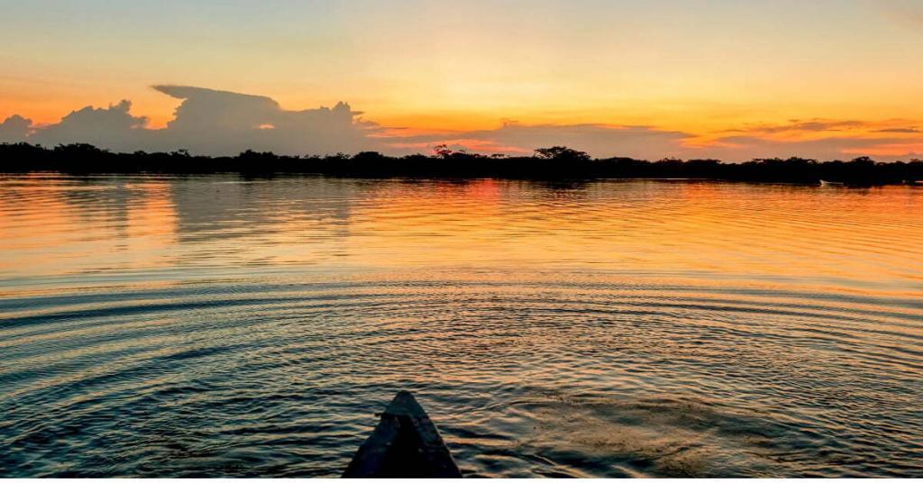 A canoe on a calm lake at sunset with vibrant orange and yellow skies reflecting on the water's surface. Silhouetted trees line the horizon.