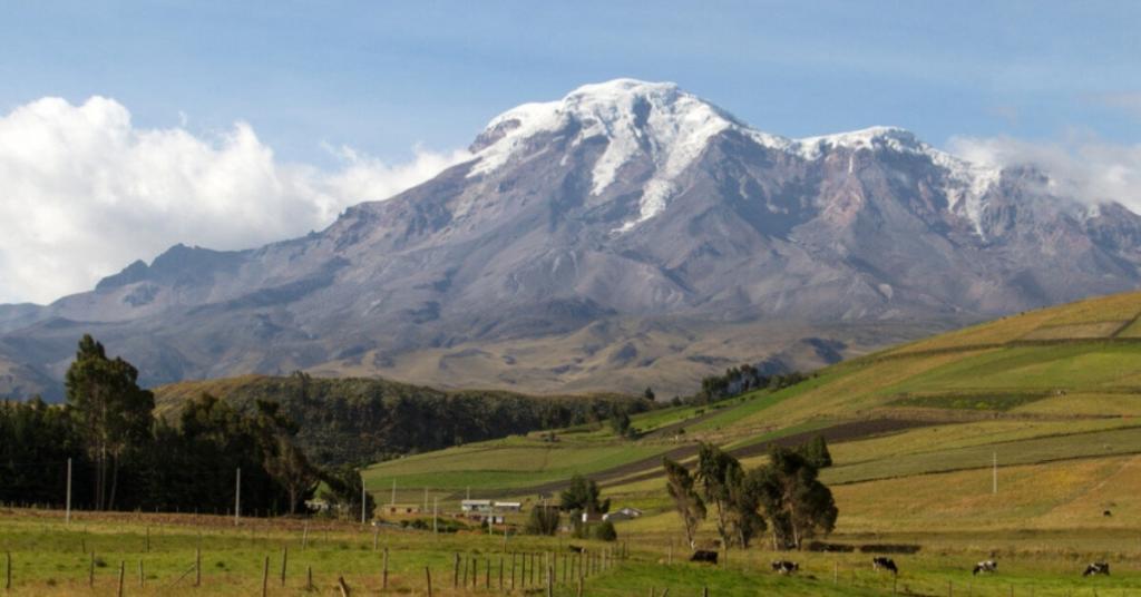 Snow-capped mountain with lush green foothills, patches of farmland, scattered trees, and grazing cows under a partly cloudy sky.