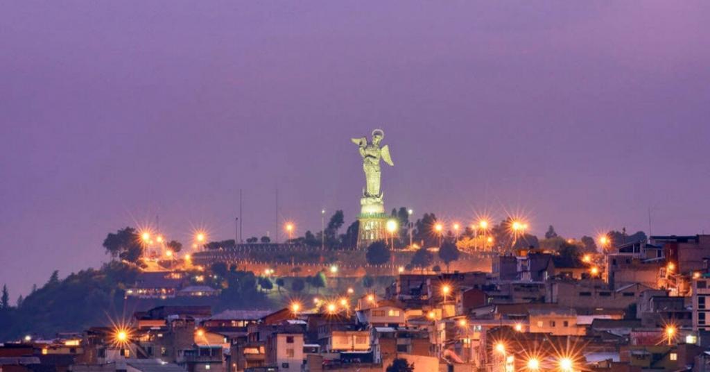 Illuminated statue of a winged figure atop a hill, surrounded by city lights and buildings at dusk.