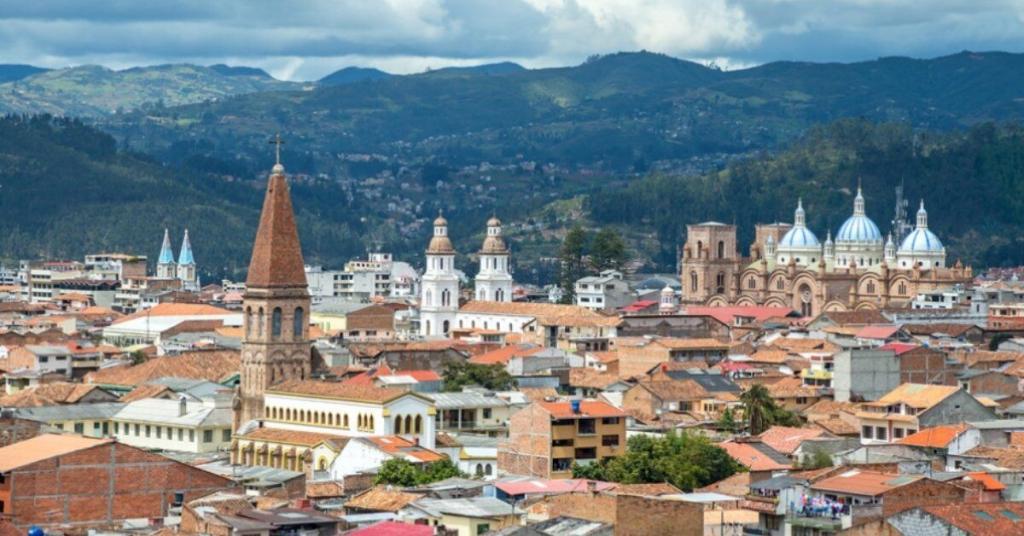 A scenic view of a city with red-tiled rooftops and several prominent churches, set against a backdrop of green mountains under a cloudy sky.