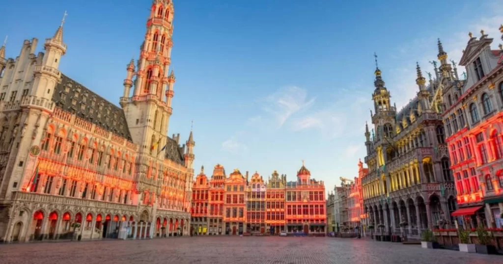 A spacious city square with ornate historic buildings under a clear sky, some illuminated with red lights.