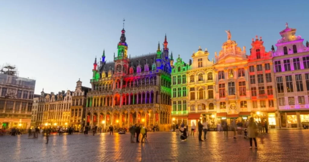 Brussels' Grand Place illuminated with multicolored lights at dusk, featuring ornate historic buildings and tourists scattered across the cobblestone square.